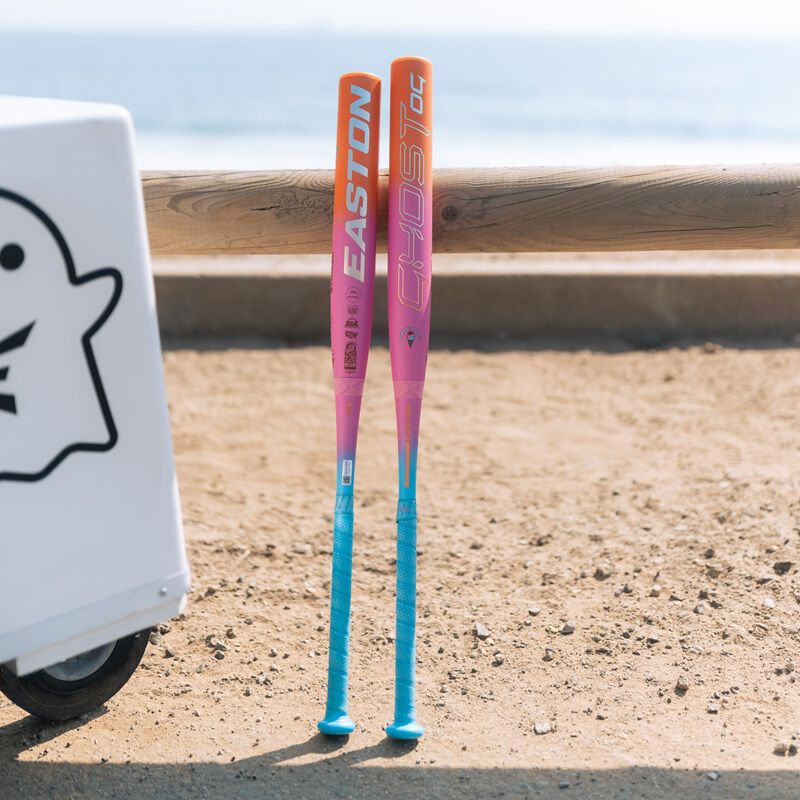 Two Easton Ghost OG Rainbow sherbet fastpitch bats leaning against a fence on the beach