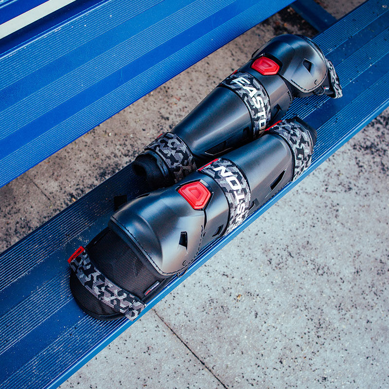 A pair of black Easton Hellcat Mojo leg guards on a bench in a dugout - SKU: HELMOLG loading=