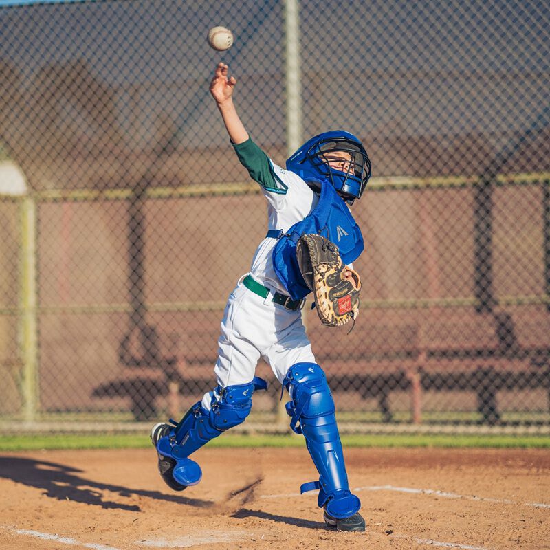 A player throwing a ball while wearing a royal Easton Black Magic 3.0 youth catcher's set