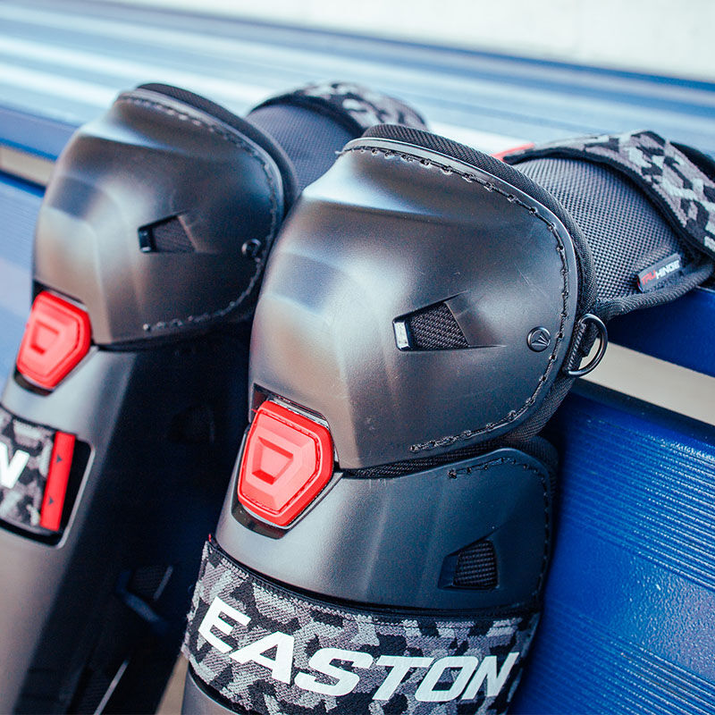 A pair of Hellcat Mojo leg guards leaning against the back of a dugout bench - SKU: HELMOLG loading=