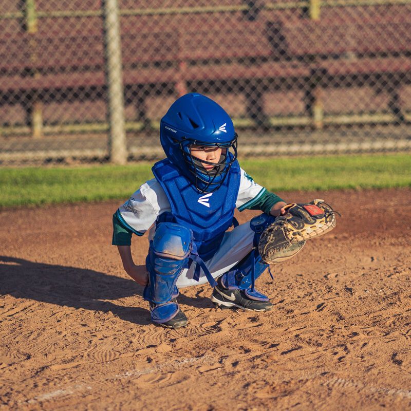 A player in his catcher's stance behind the plate wearing a royal Easton Black Magic 3.0 youth catcher's set