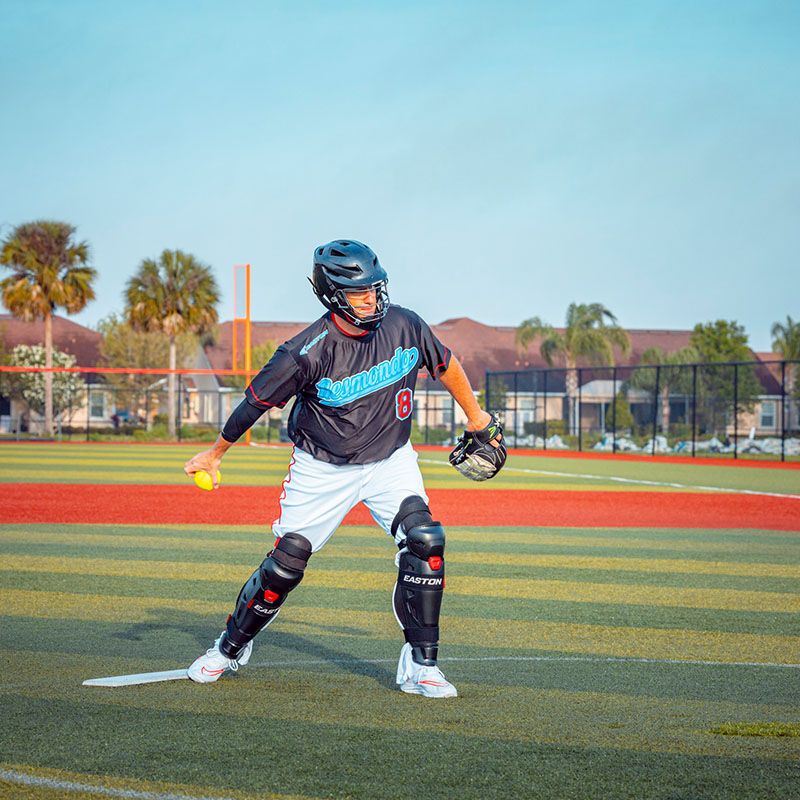 A slowpitch player pitching a ball wearing a black Easton Hellcat slowpitch fielding helmet and leg guards - SKU: EHCATH