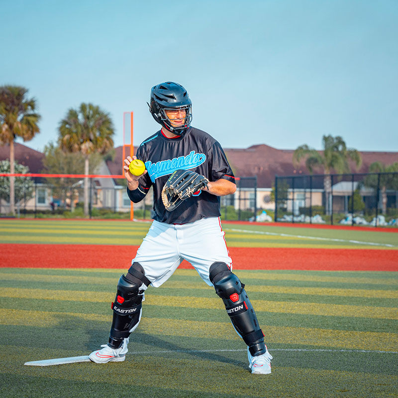 A pitcher on the mound wearing a black Easton Hellcat fielding helmet and leg guards - SKU: EHCATH loading=