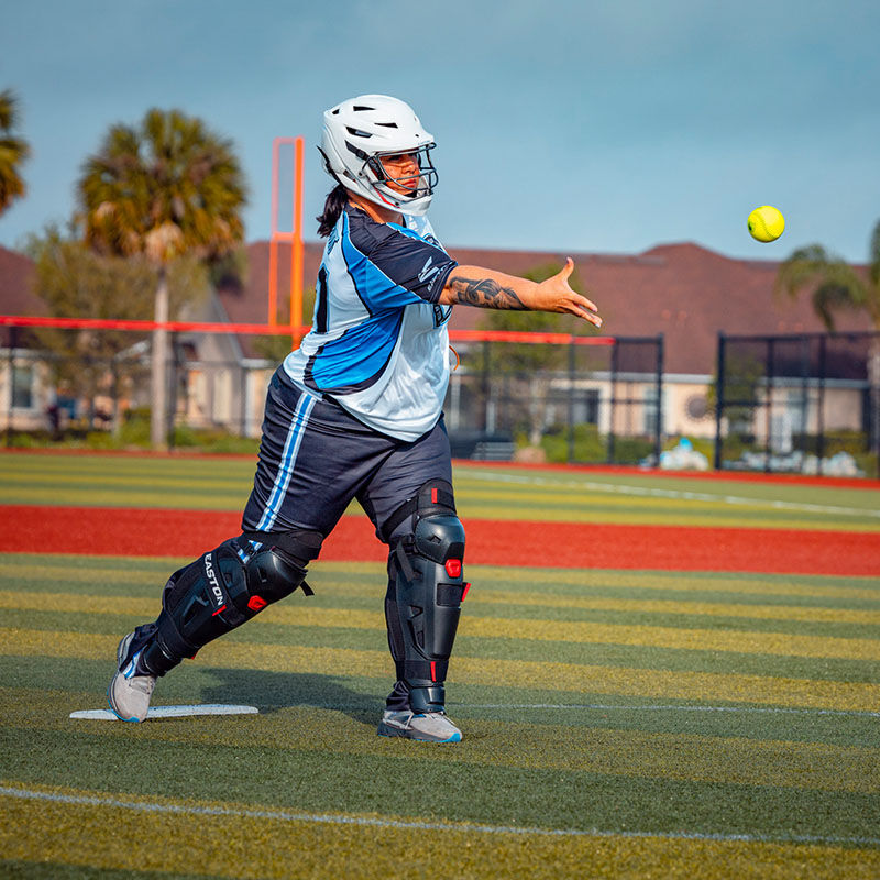A female pitcher throwing a pitch wearing a white Hellcat helmet and black Hellcat leg guards - SKU: EHCATH loading=